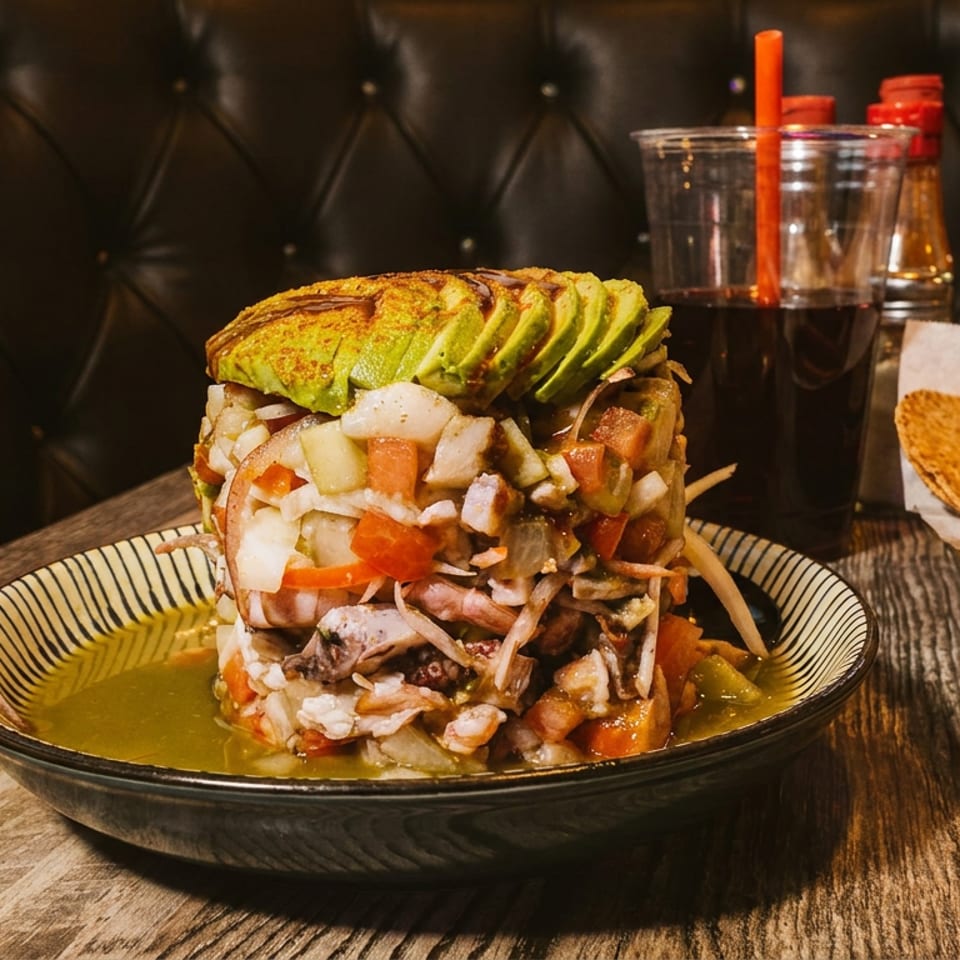 A tall ceviche tower with avocado slices, tortilla chips, and drinks on a wooden table.