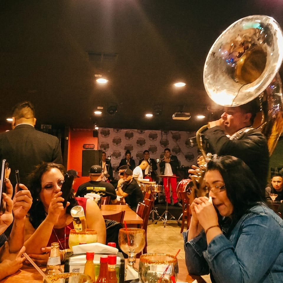 A band playing live music in a restaurant with patrons at tables, some recording on phones.