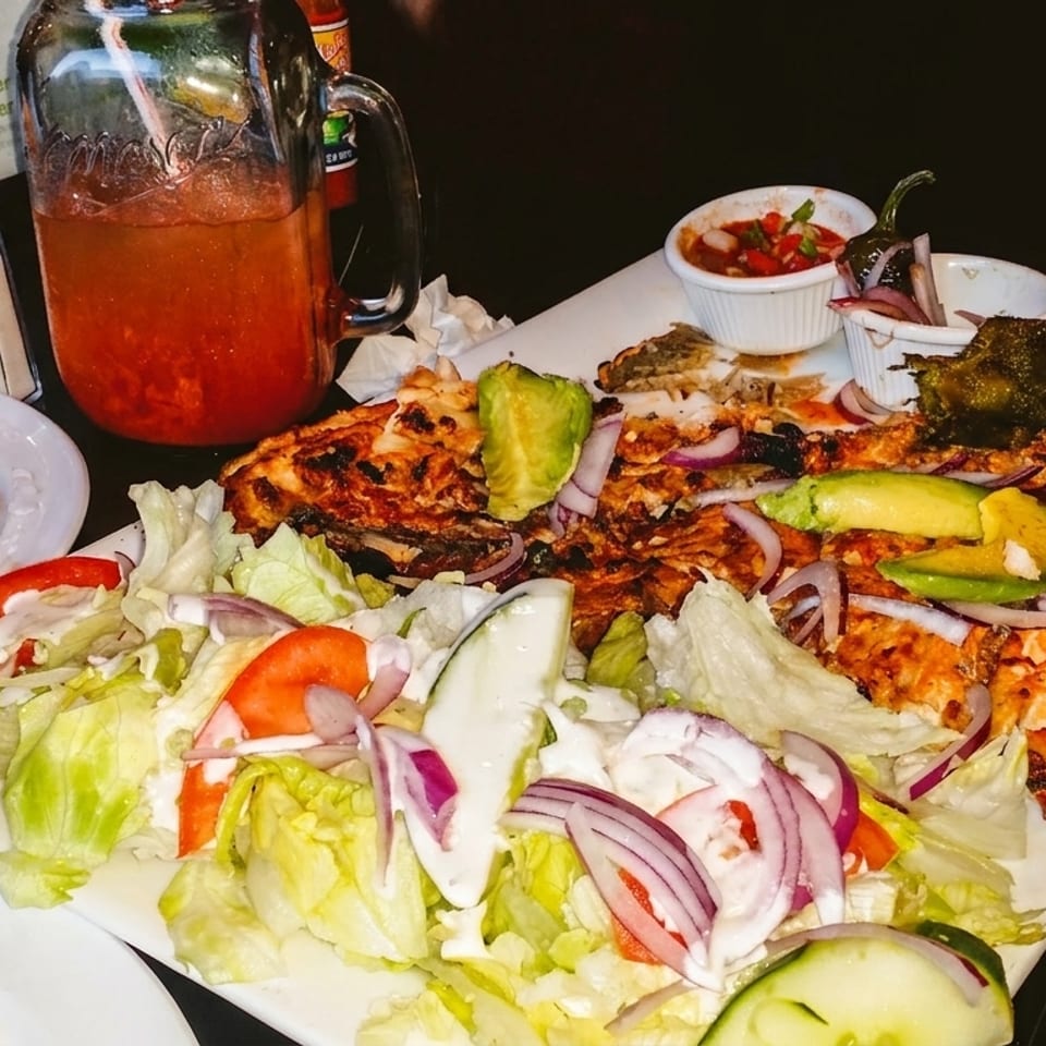 Grilled whole fish with avocado, red onion, and salad, alongside oysters and a drink.