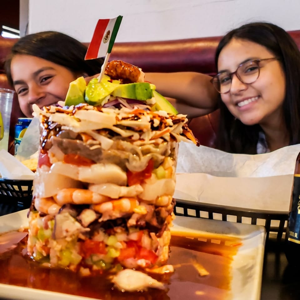 A towering seafood cocktail with a Mexican flag, beer, and two smiling women in a restaurant.