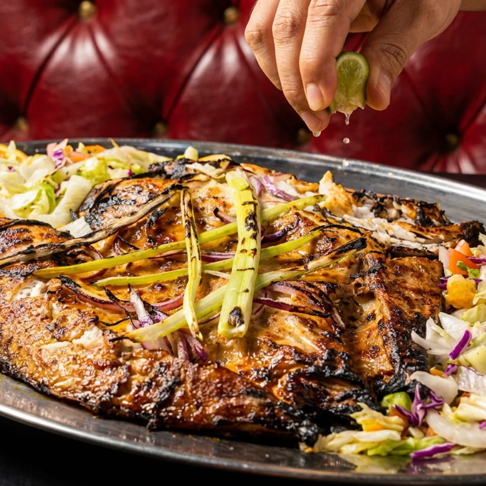 A hand squeezes lime over a large grilled fish with salad and orange slices on a platter.
