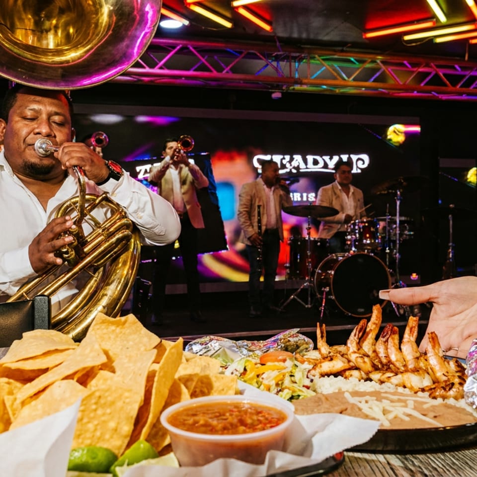 Man playing a tuba on stage with a band, with chips, salsa, and shrimp on a table.